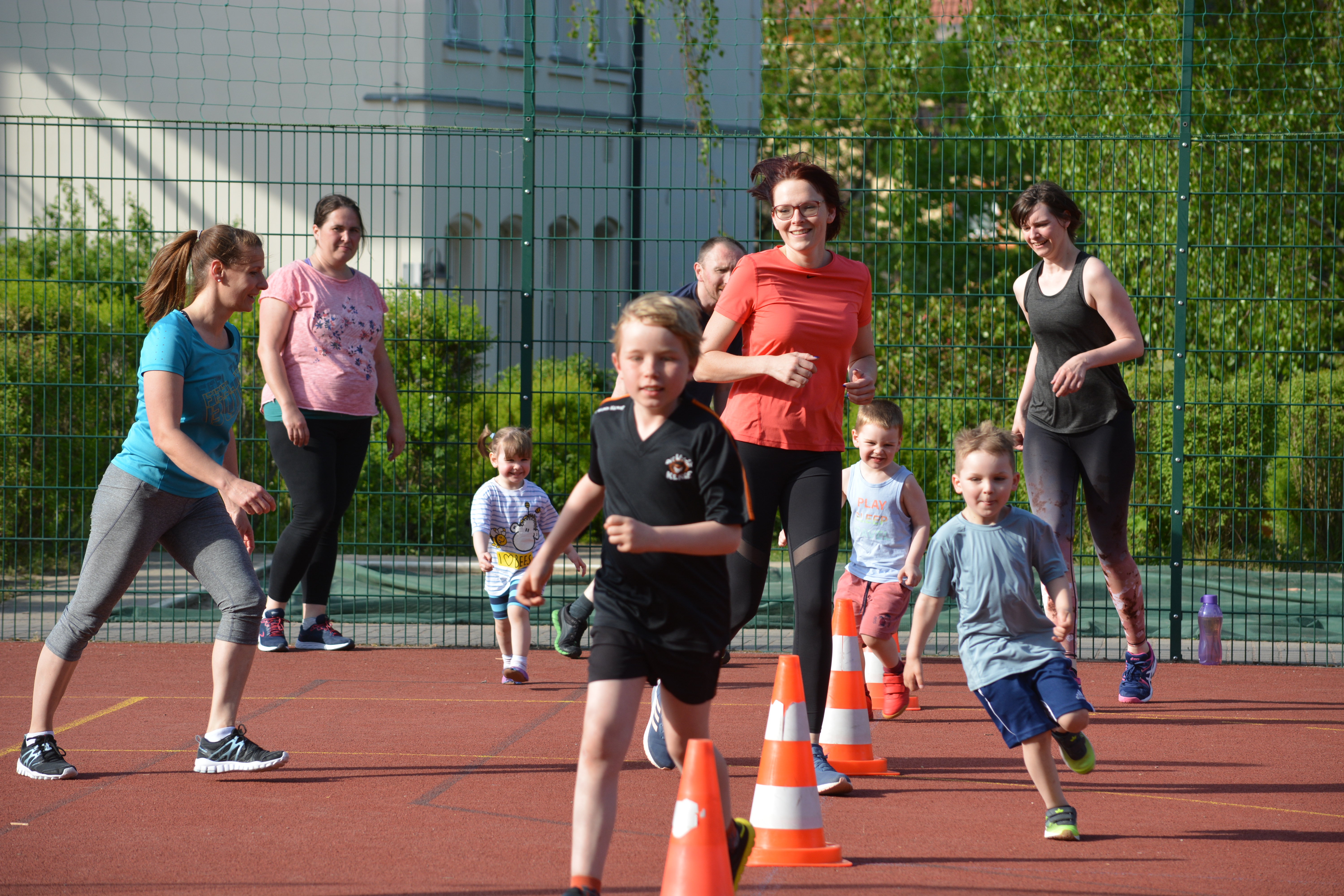 mehre Kinder und Eltern in sportlichen Anziehsachen rennen durch einen Slalom-Parcours aus Verkehrskegeln. Sie befinden sich draußen bei sonnigem Wetter auf einer Außensportanlage mit rotem Kunststoffboden.