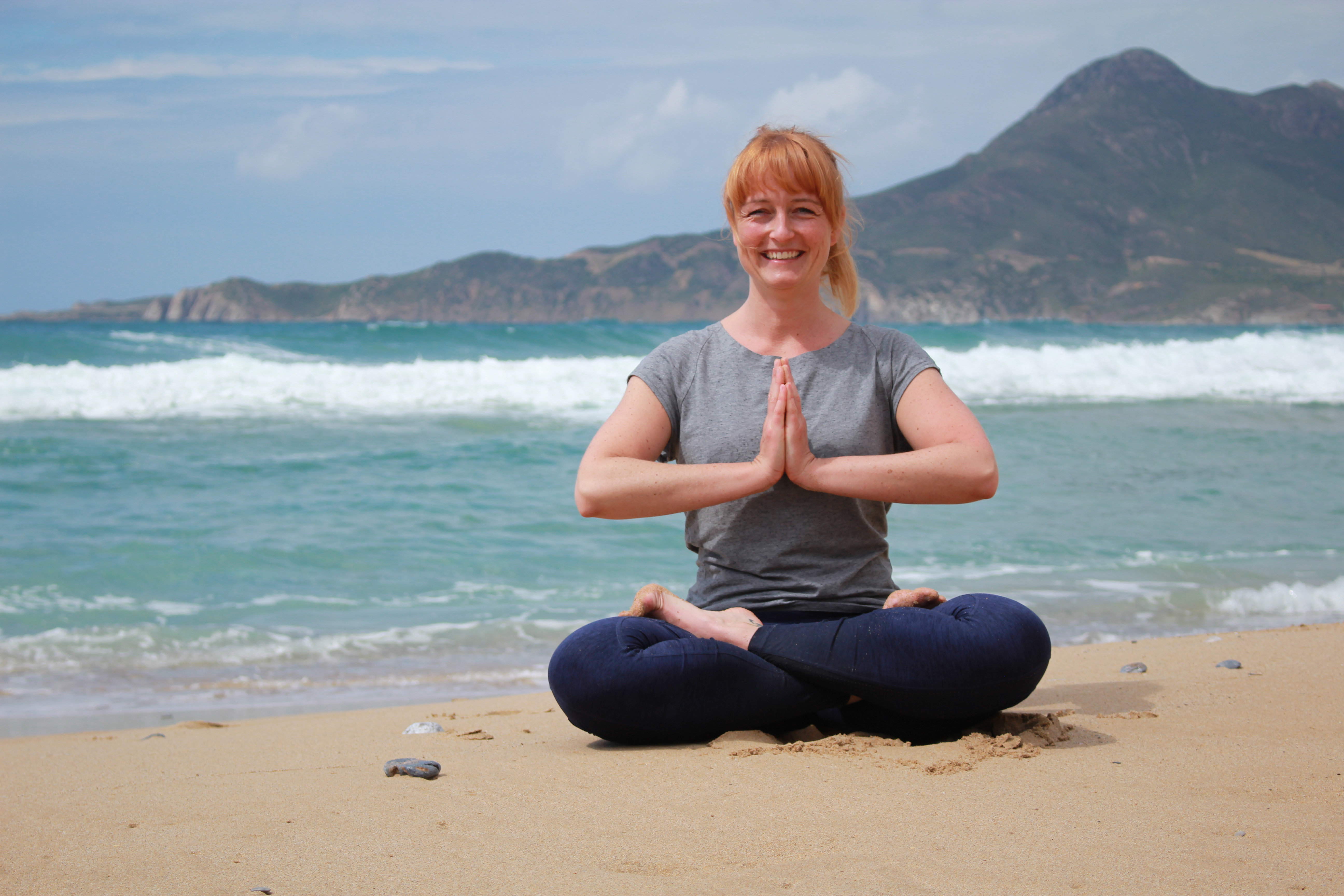 Eine lächelnde junge Frau mit roten Haaren sitzt in lockeren Anziehsachen im Yogi-Sitz am Sandstrand im Sand. Im Hintergrund ist eine Meereswelle und das andere Ufer in Form eines mittelhohen Berges, der sich über die gesamte Bildbreite erstreckt, zu sehen